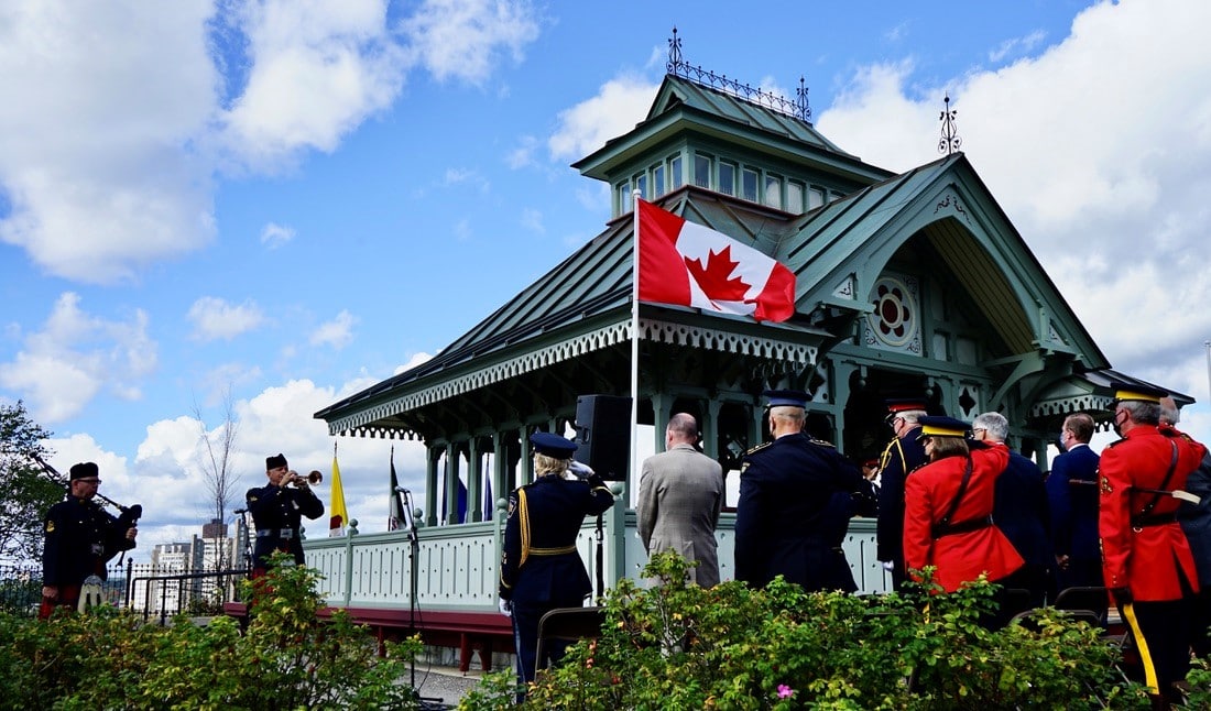 Photo of people standing in front of a pavilion on a cloudy day. To the left, a bagpiper is playing.