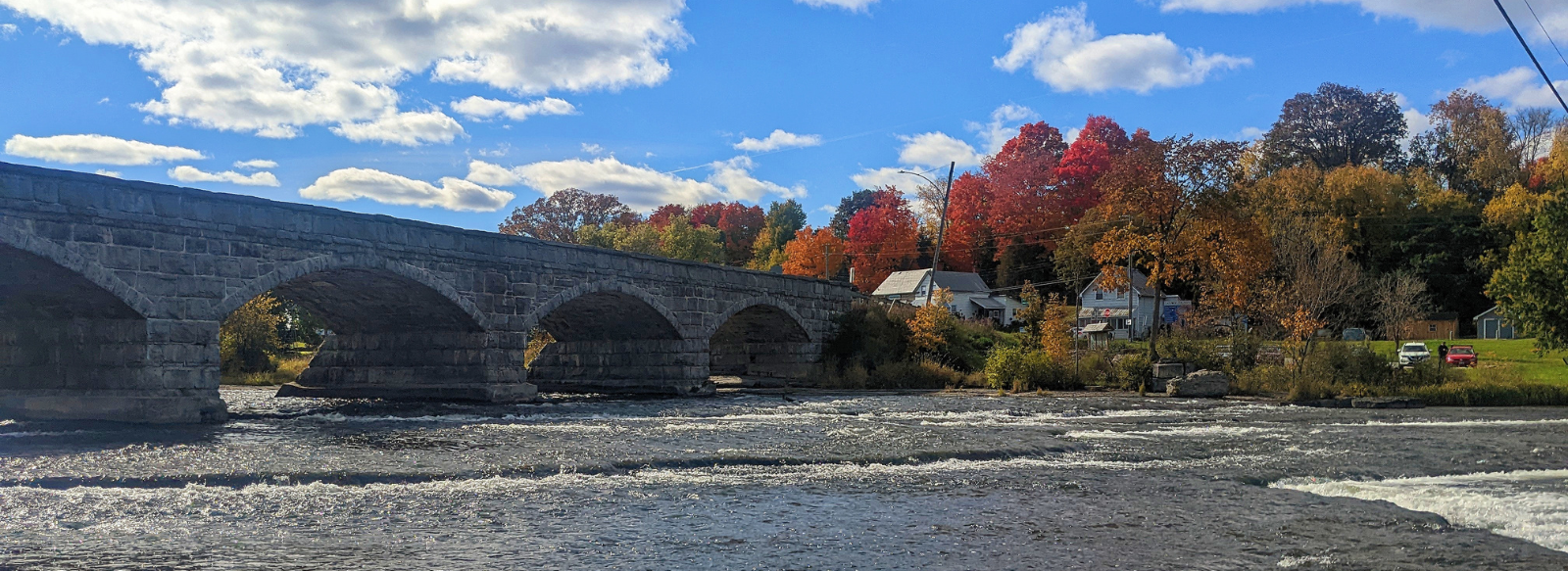 A stone bridge crosses a river with trees displaying fall colours