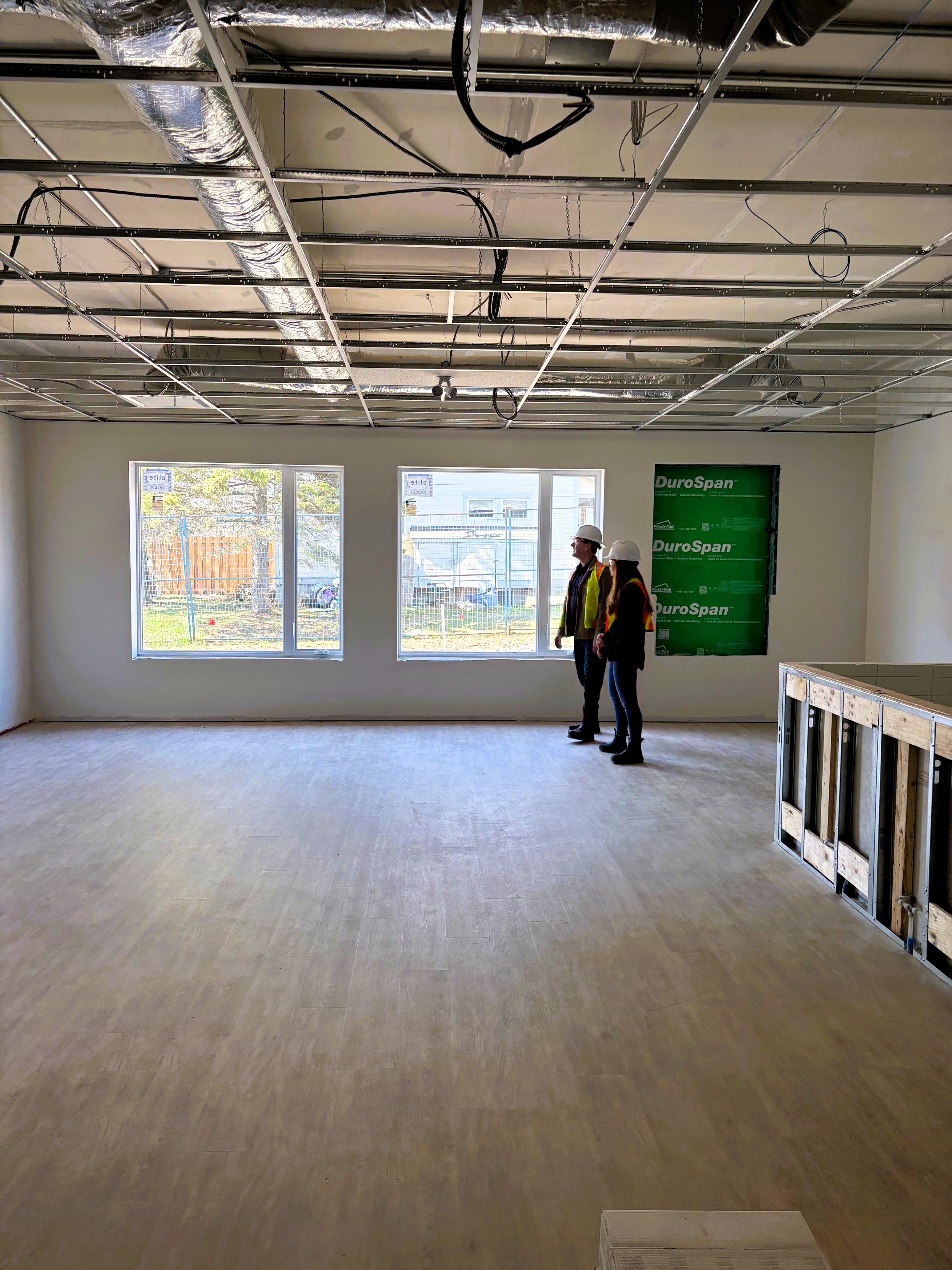 Two people in hard hats and orange and yellow construction vests look at the inside of a building under construction