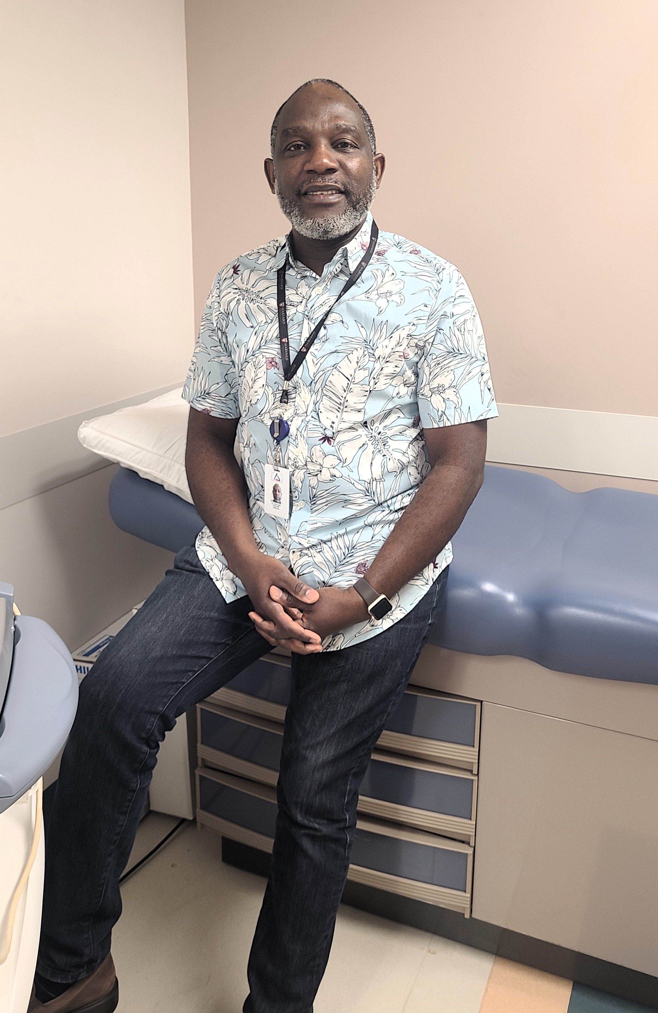 Older Black man in a blue Hawaiian print shirt stands against a hospital bed in a clinic room